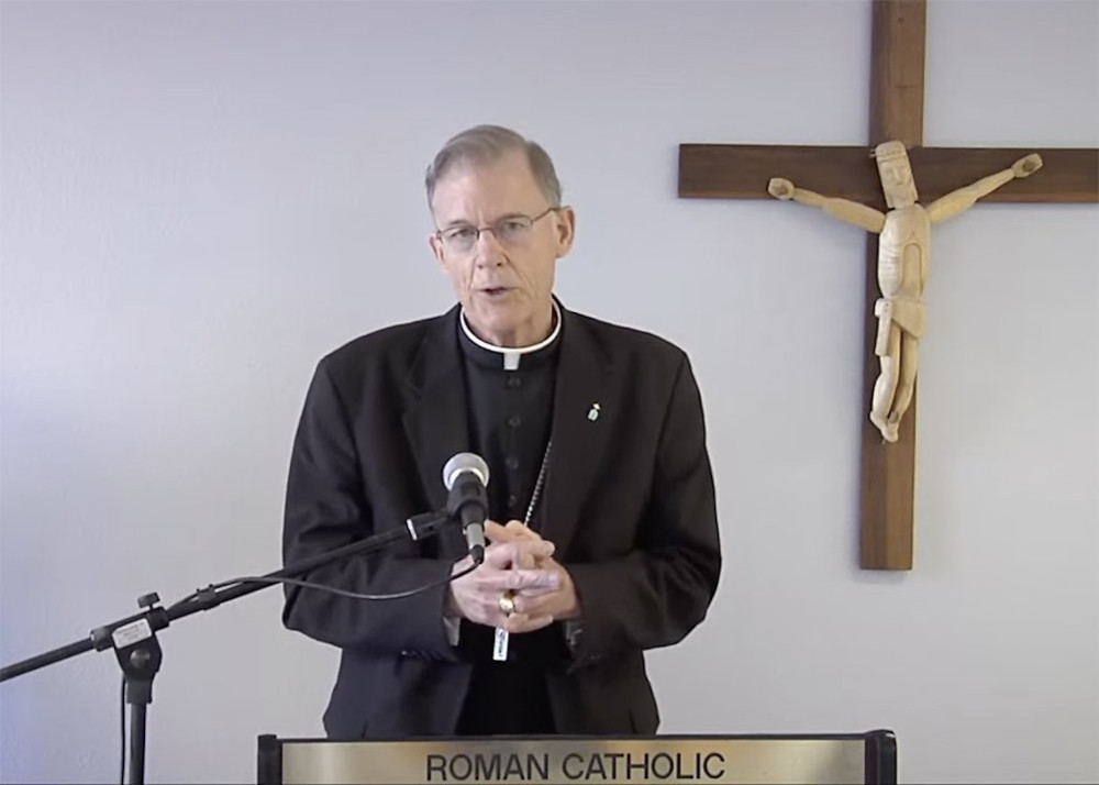 A older, white cleric stands at a lectern with a microphone in front of a crucifix