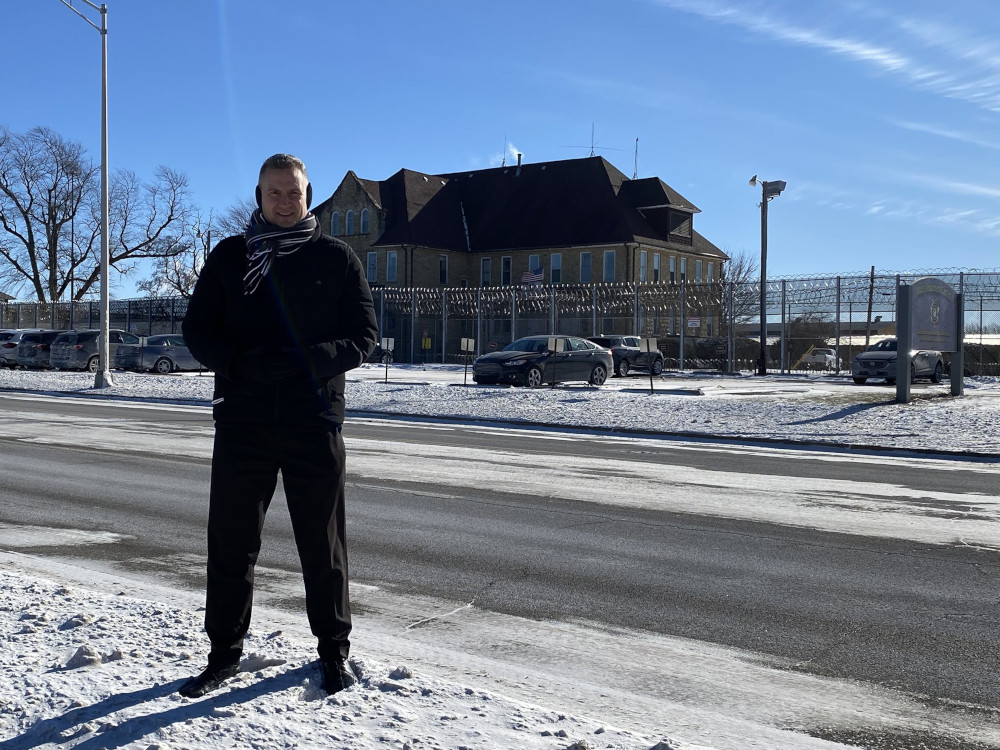 A white man wearing a black coat stands in the snow on the side of a road in front of a building