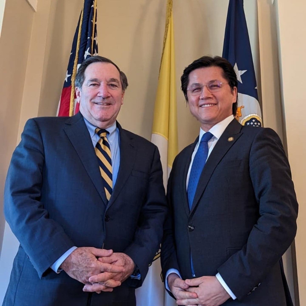 An older white man in a suit stands next to a Brown-skinned man in a suit