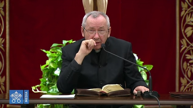 A priest in a black cassock speaks into a microphone in front of a burgundy backdrop