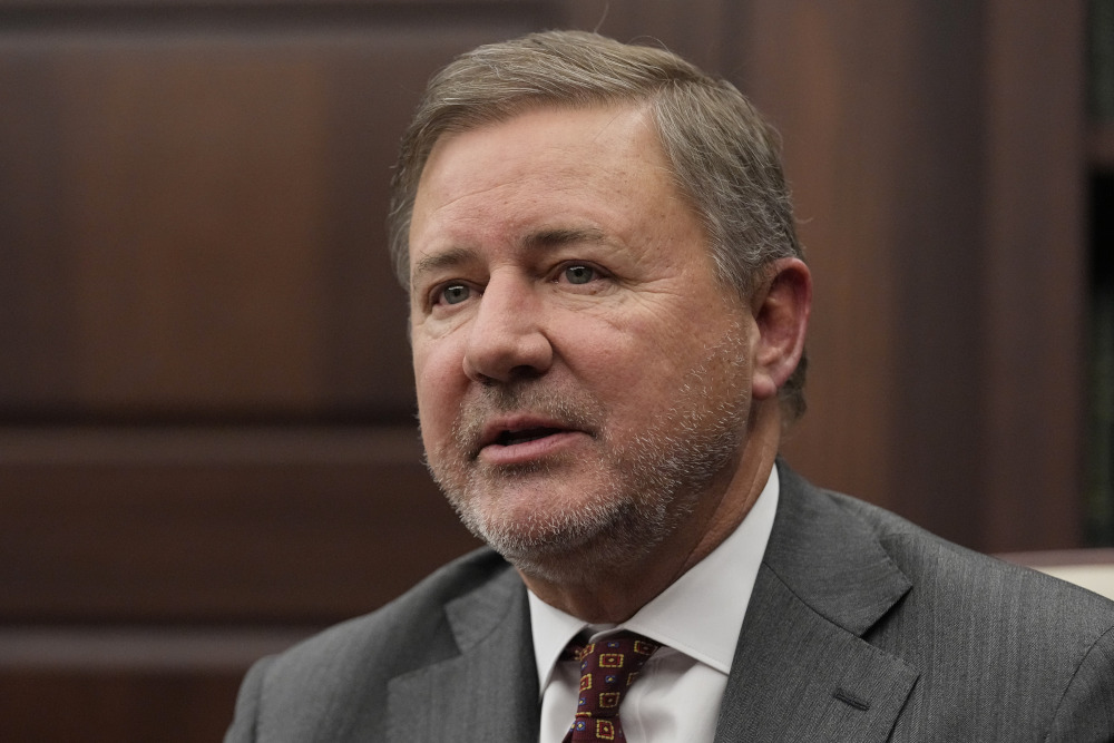 A white man with gray stubble wearing a suit and tie looks into the camera