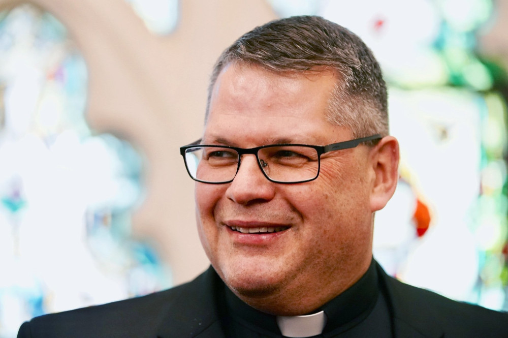 A white man with a short haircut wears glasses and a clerical collar and smiles.