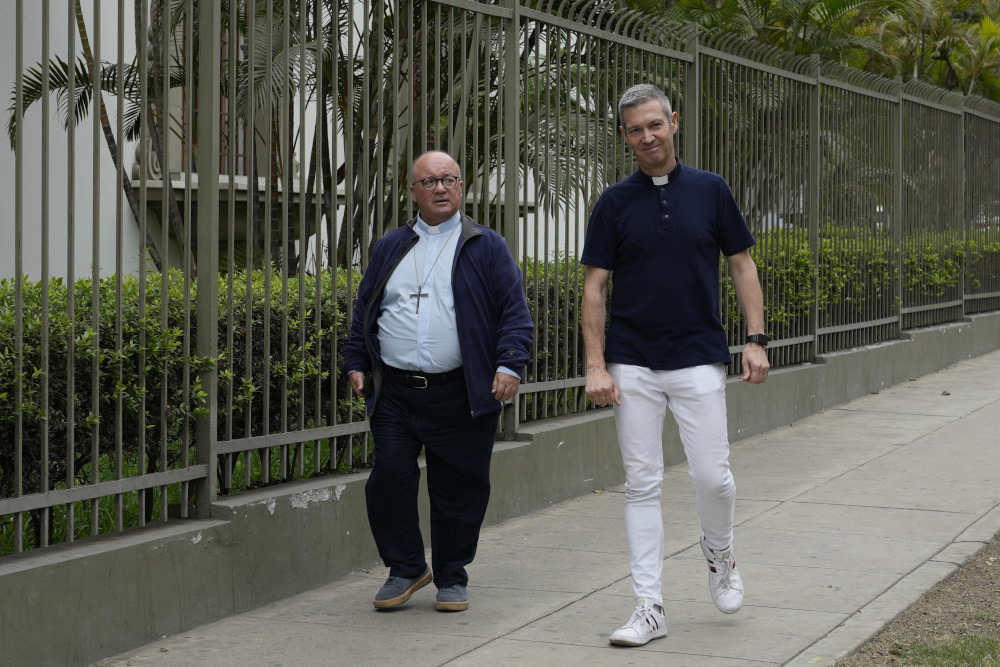 A bald man wearing a light blue shirt, clerical collar and pectoral cross walks next to a gray-haired man with a short-sleeved dark shirt with a clerical collar down a sidewalk