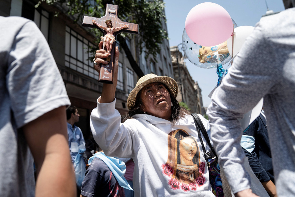 A woman wearing a shirt with the Virgin Mary on it holds a crucifix above her head while in a crowd