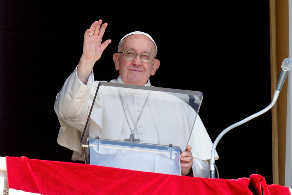 Pope Francis raises his hand to wave as he stands behind a clear lectern. There is a red tapestry over his window ledge.