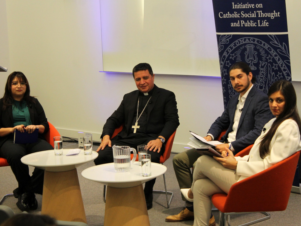 Four people, including two women on the ends, a man wearing a clerical collar and jacket and another man wearing a jacket in the middle, sit in chairs around two round tables