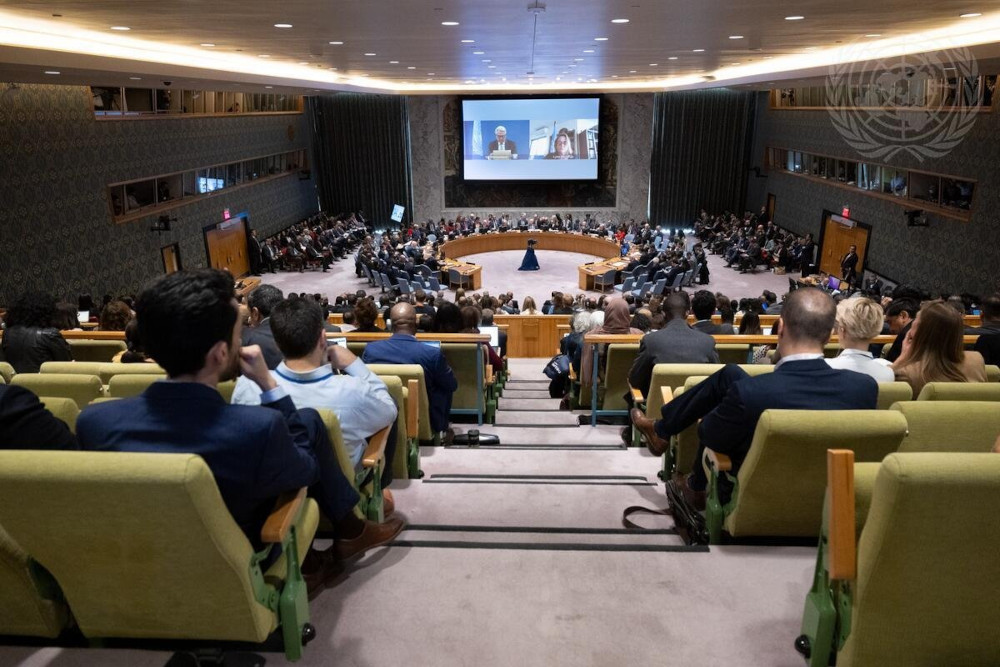 People, mostly men in Western business attire sit in yellow chairs looking down on circular table with many people around it