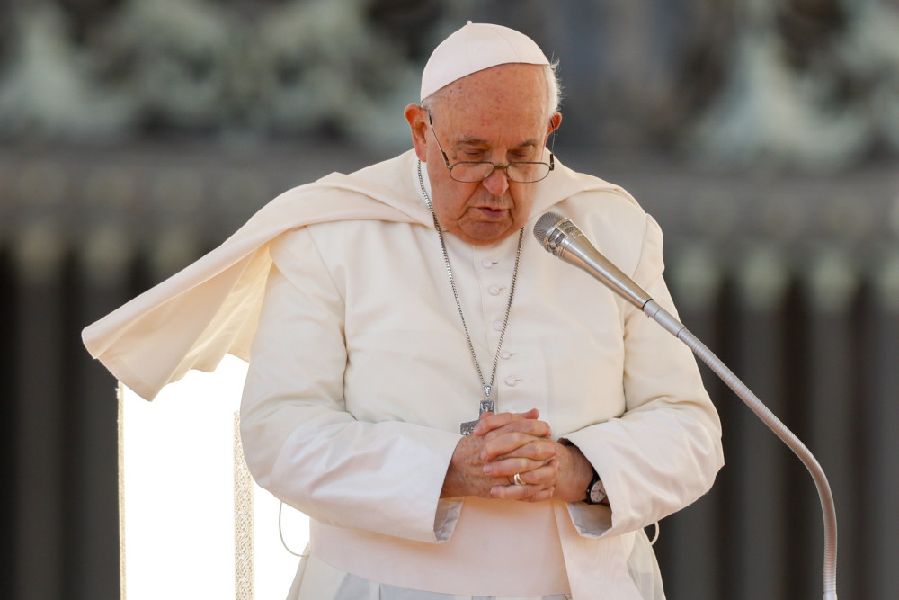 Pope Francis bows his head and folds his hands as he stands in front of a microphone and his mozzetta is blown to the side.