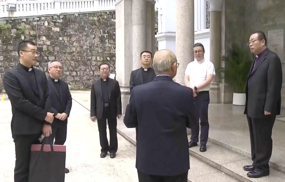 A group of men, mostly in clerical collars, stand in a circle outside a stone building