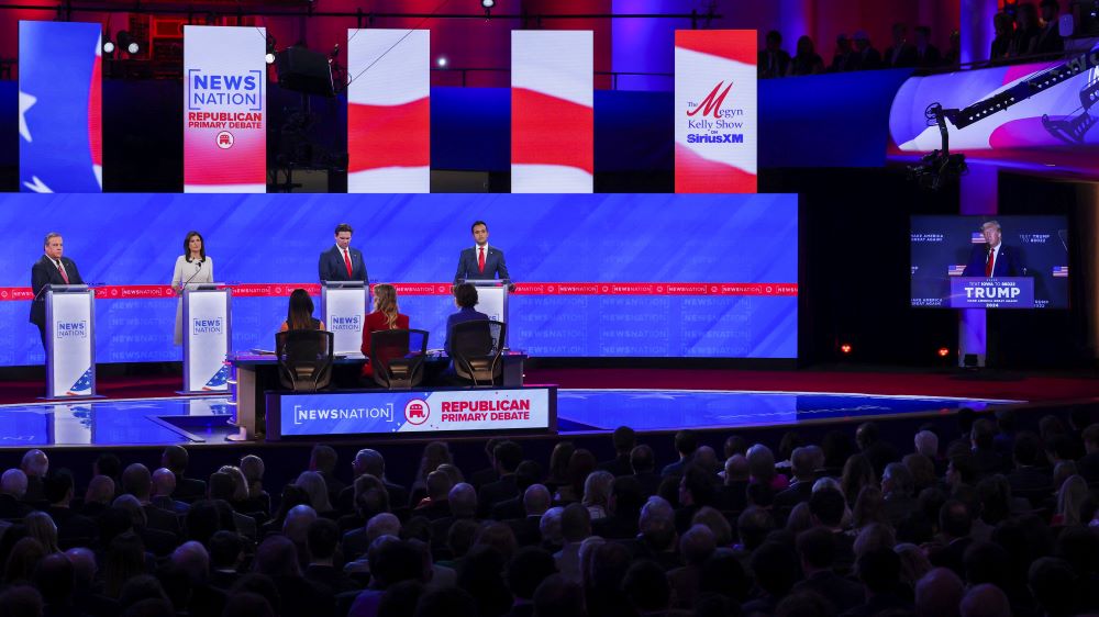 Four presidential candidates on stage for debate, while screen to the right shows former U.S. President Donald Trump