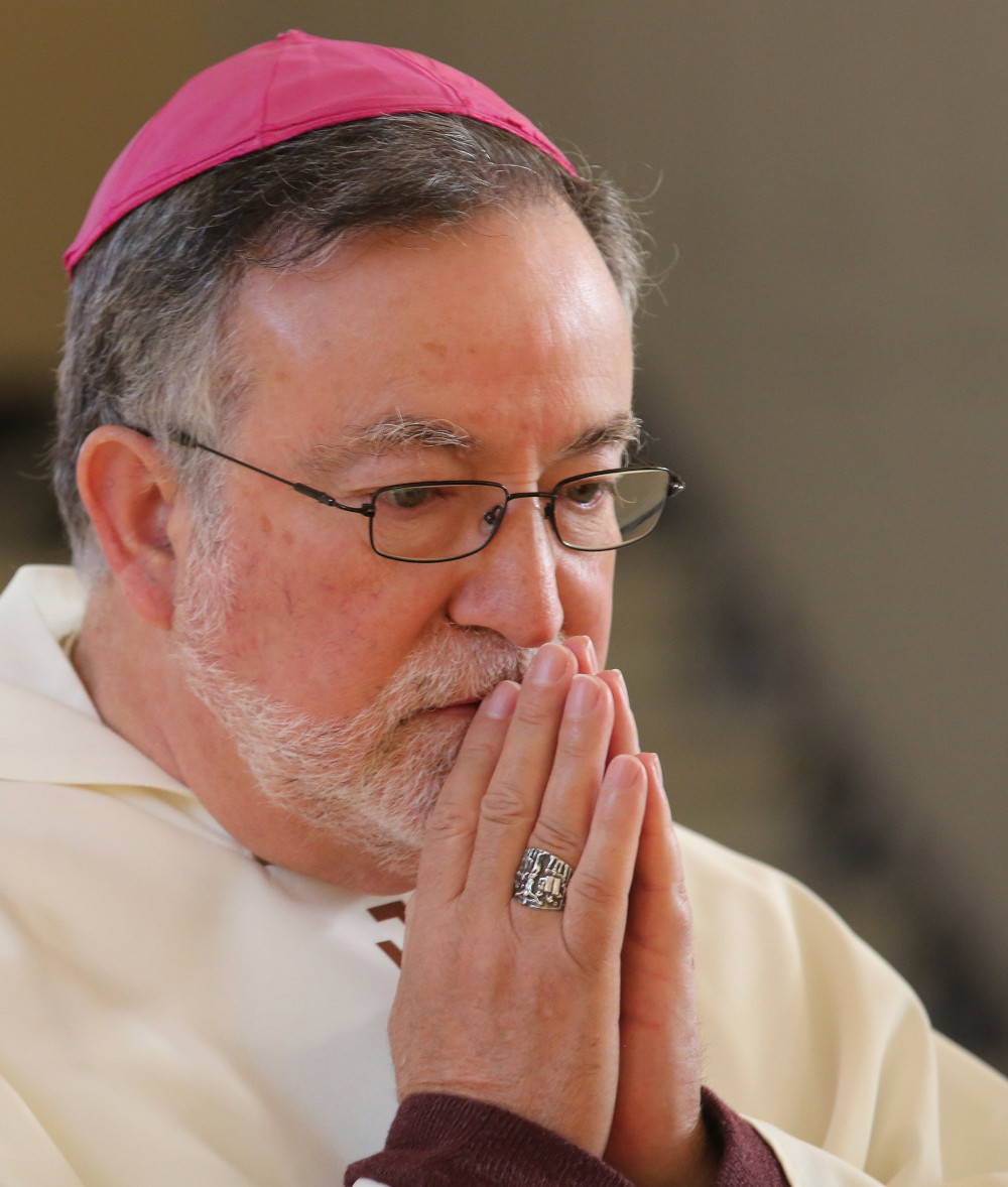 A man wearing a violet zucchetto holds his hands palms together in front of his mouth