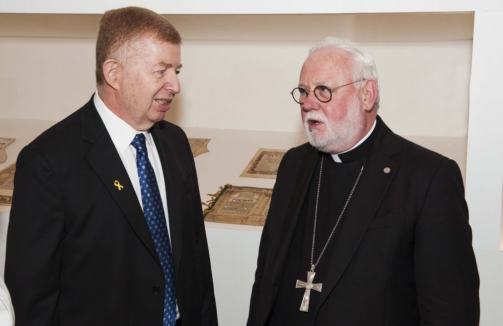 The two men stand facing each other, talking in front of a museum display case