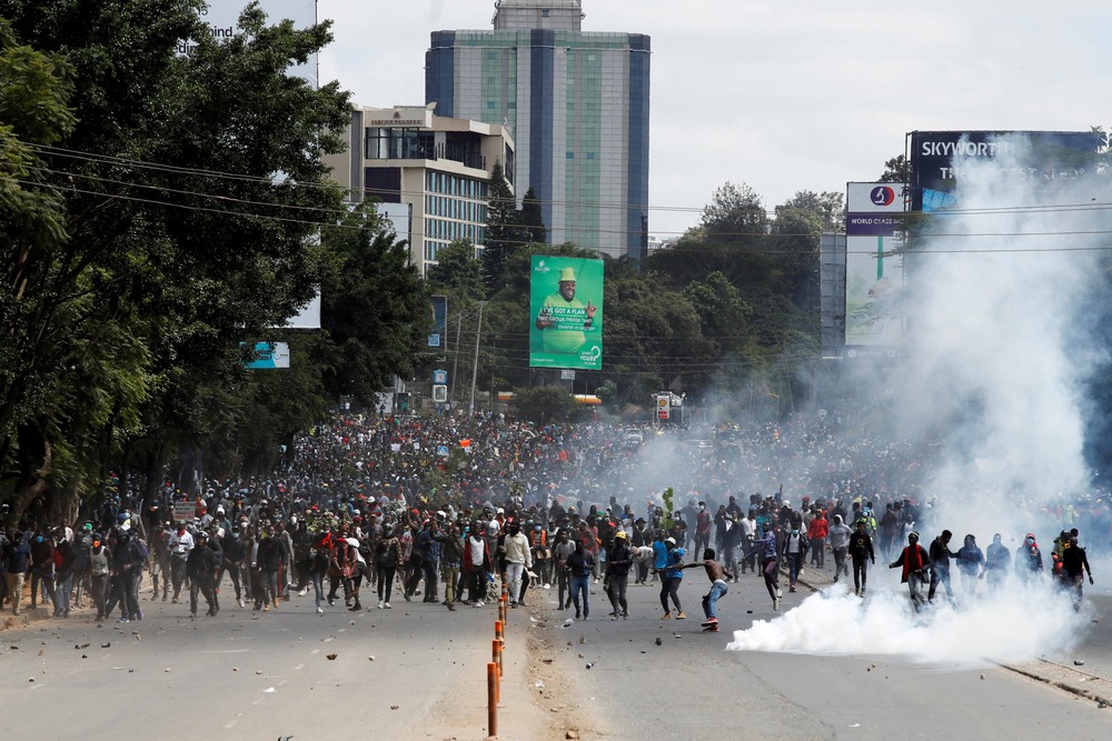 Enormous assembly marches through wide boulevard as smoke rises from street.