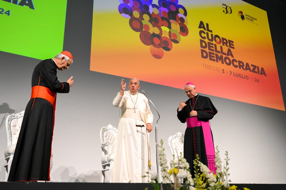 Pope standing on stage with two prelates, raises hand in gesture. 