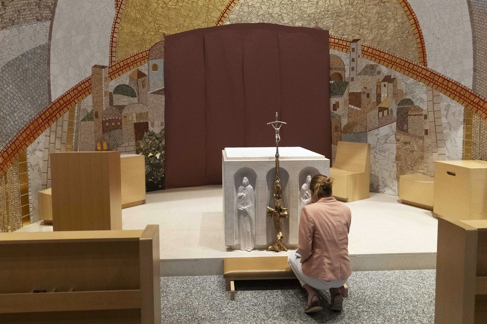 Woman kneels at small altar, behind altar are Rupnik's mosaics. 