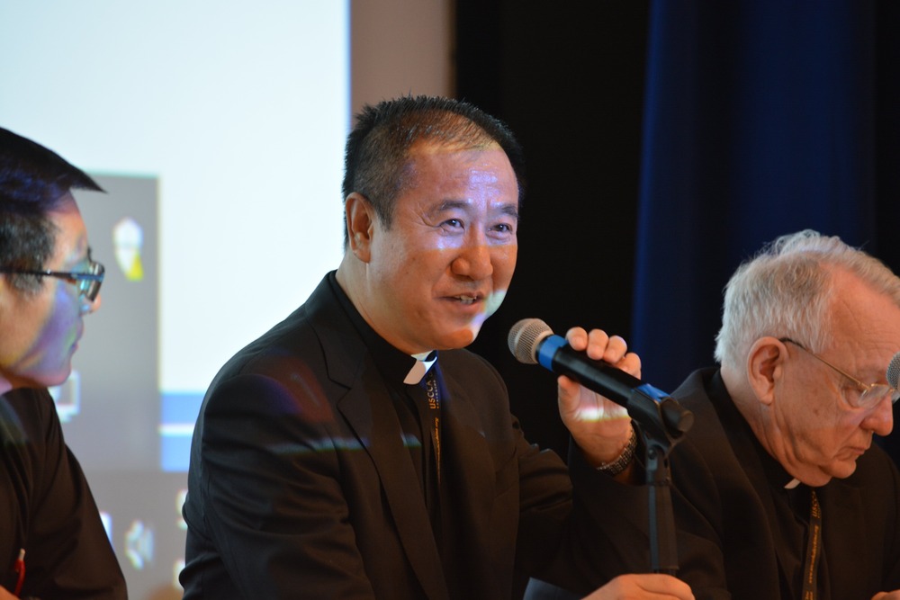 Bishop Pei sits at panel, wearing black clericals, and speaking into microphone. 