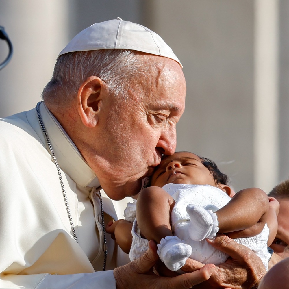 Close-up, square photo of Pope kissing a sleeping infant in the warm glow of the sun. 