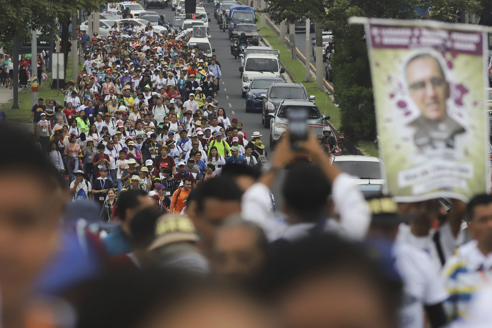 Large banner of Romero carried above long assembly of people marching down street. 