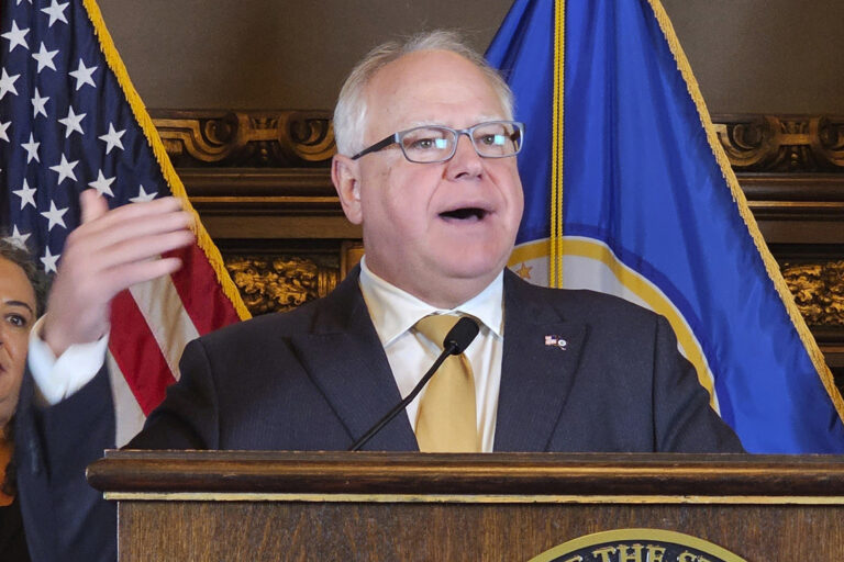 Gov. Walz stands at podium speaking, behind are US and Minnesota flags. 