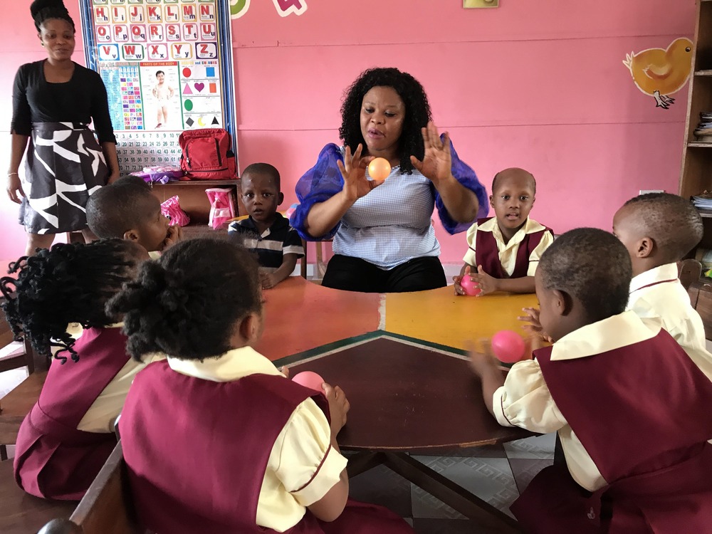 Teacher sits at table with students, holding a yellow sphere. 