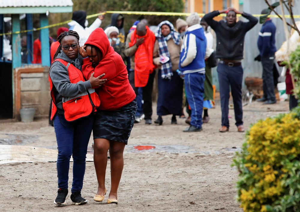 A crowd of mourners; two women walk away towards the camera, one holds the other as he cries. 