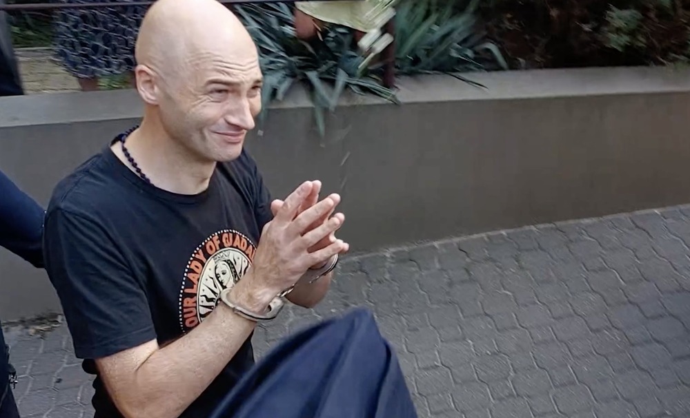 Olszewski smiles with hands folded in prayer; his head is shaven, and he is thin.