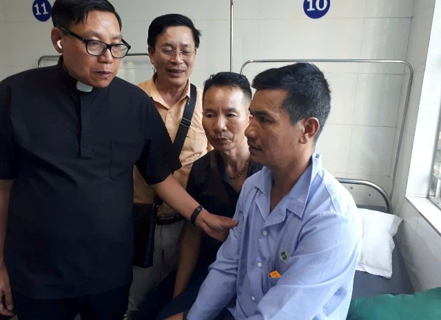 A man sits on hospital bed; next to him are the priest and two others.