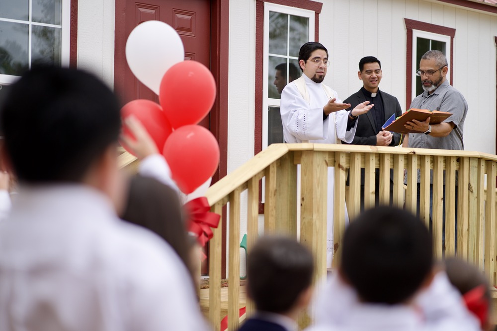 De Leon and others stand on the wooden porch of a prefab building saying a blessing; balloons and audience members frame shot.