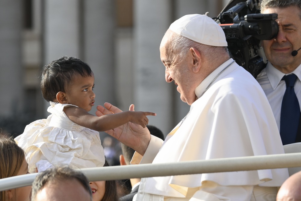 Small child holds out arm as Pope smiles.