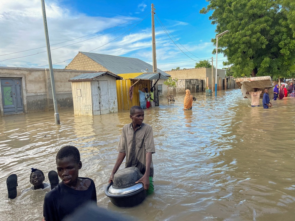 Street filled with waist-high water; foregrounded are three young men carrying items as they wade through water. 