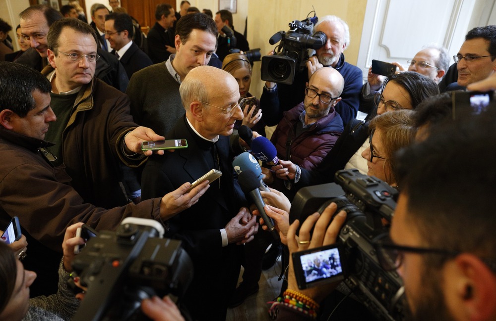 Overhead view of prelate surrounded by journalists extending microphones and cell phones.