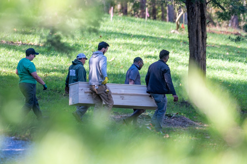 Seen through a frame of leaves, are five people, four of whom carry a casket.