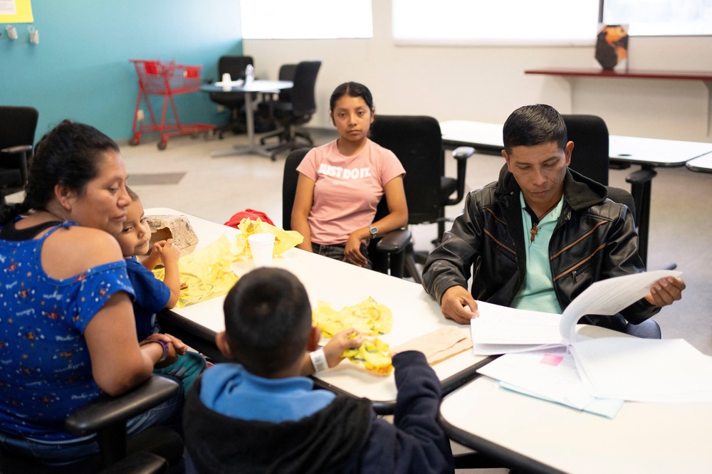 Family sits around table with trays of food in what looks like a classroom.