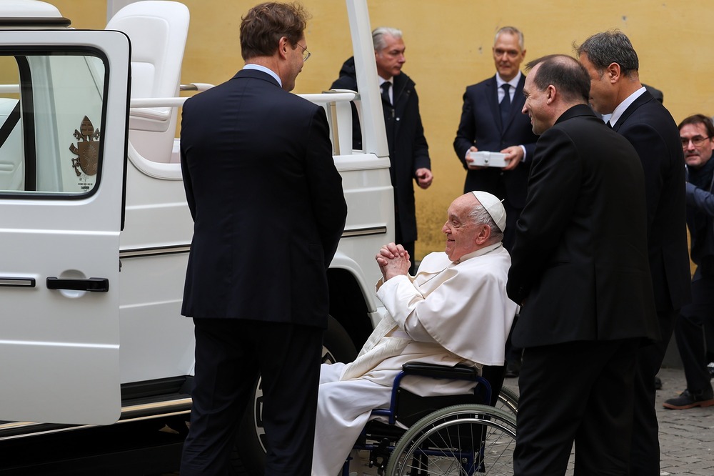 Francis seated in wheelchair, and surrounded by Mercedes reps, looks at new popemobile.