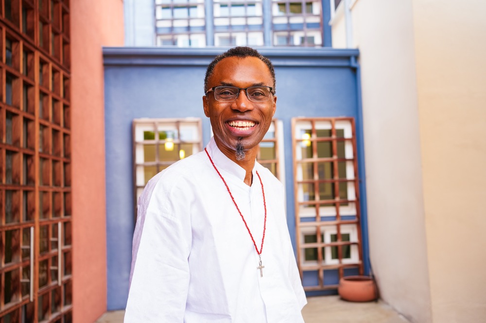 The priest wears white clothing and faces camera smiling.