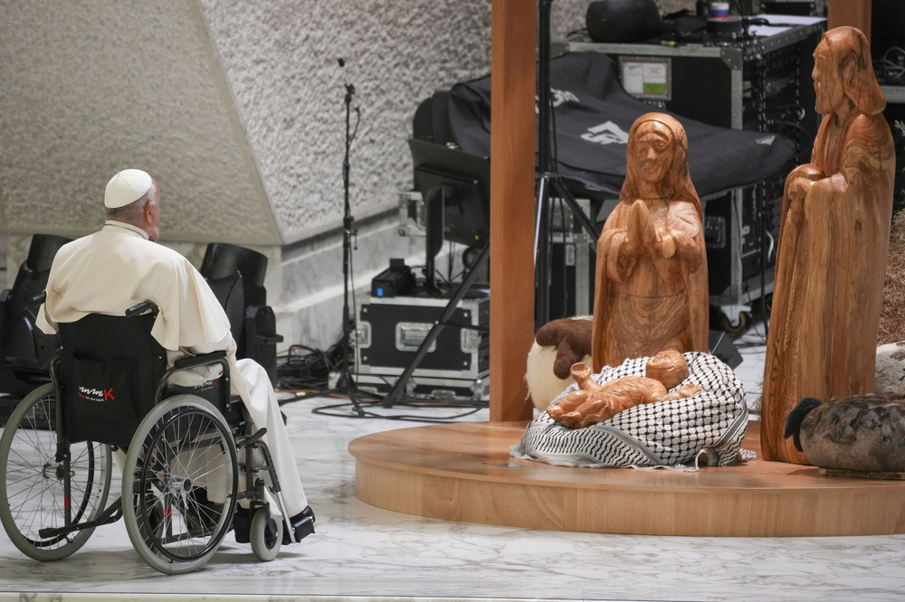 Pope Francis in wheelchair sits before wooden nativity scene with keffiyeh.