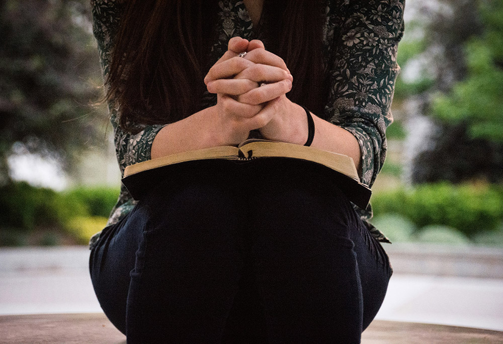 A woman sits outside with her hands folded on top of an open book, possibly a bible. (Unsplash/Olivia Snow)