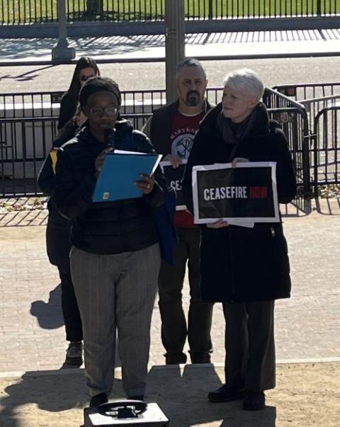 Four people stand together, one holding a sign that says, "ceasefire." A woman speaks into a microphone.