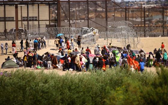 Many people with tents and umbrellas stand on desert land in front of barbed wire structures in front of the rust-colored U.S. border wall