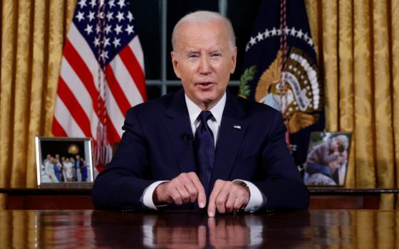 President Biden wearing a jacket and tie sits at a desk in the Oval Office with the American flag behind him