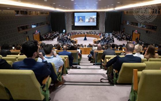 People, mostly men in Western business attire sit in yellow chairs looking down on circular table with many people around it