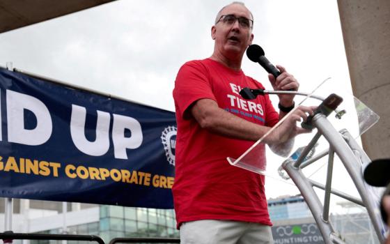 A white man wearing glasses wears a red t-shirt and speaks into a microphone from behind a podium