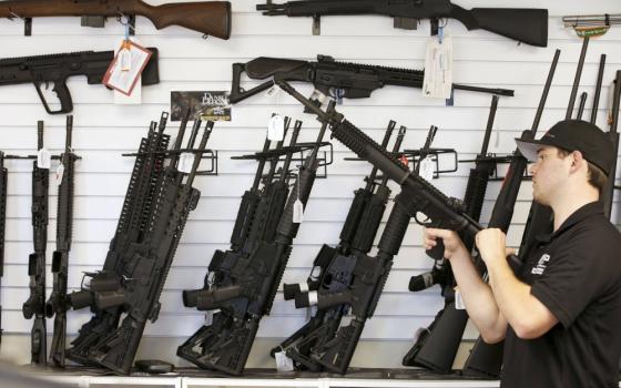 A white man wearing a ball cap holds an AR-15 against his chest. On the wall behind him are many guns, including other AR-15s.