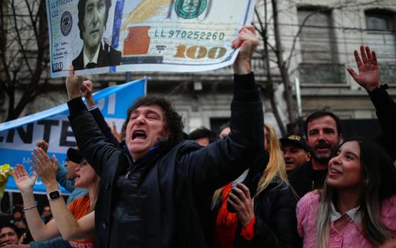 A white man with black hair and sideburns holds a $100 U.S. bill with his face on it, while in a crowd.