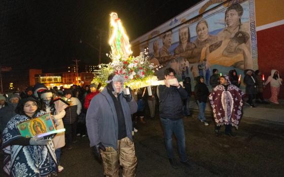 In the center of a crowd outdoors, brown men carry an illuminated Virgen de Guadalupe statue whose base is surrounded by flowers