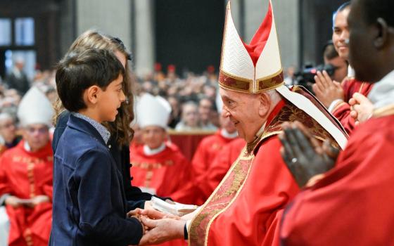 Pope Francis, seated and vested in red, bends and smiles tenderly to two small children. 