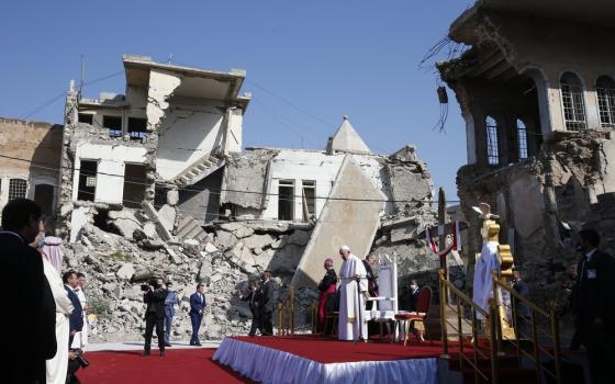 Pope Francis stands on red and white dais with rubble of bombed buildings directly behind