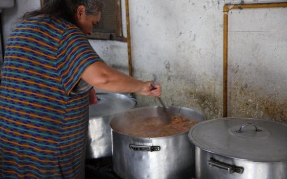 Woman bends over stove stirring food in large aluminum pots