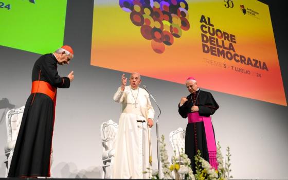 Pope standing on stage with two prelates, raises hand in gesture. 