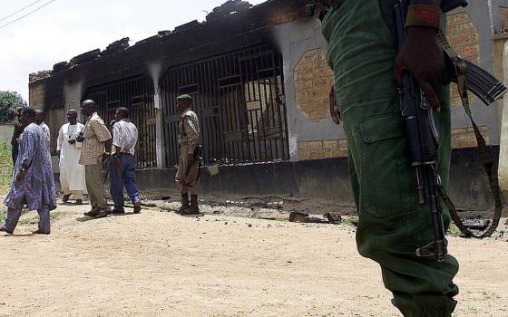 Armed police stand outside burnt shell of building; civilians come and go. 